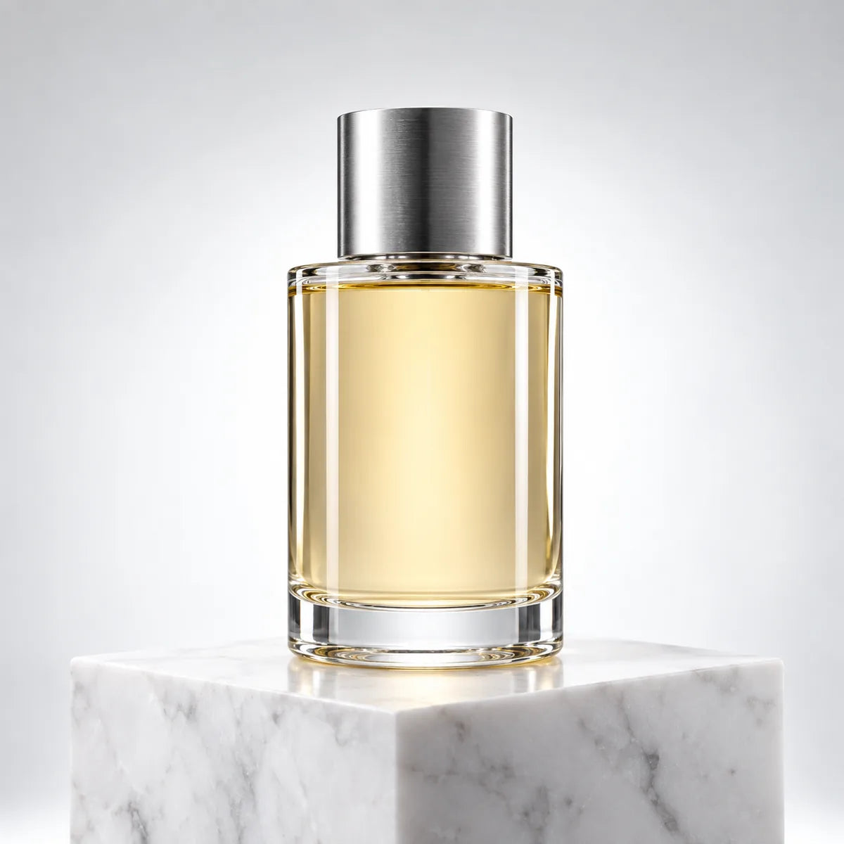 A clear cylindrical glass perfume bottle with golden liquid and a silver cap on a marble cube