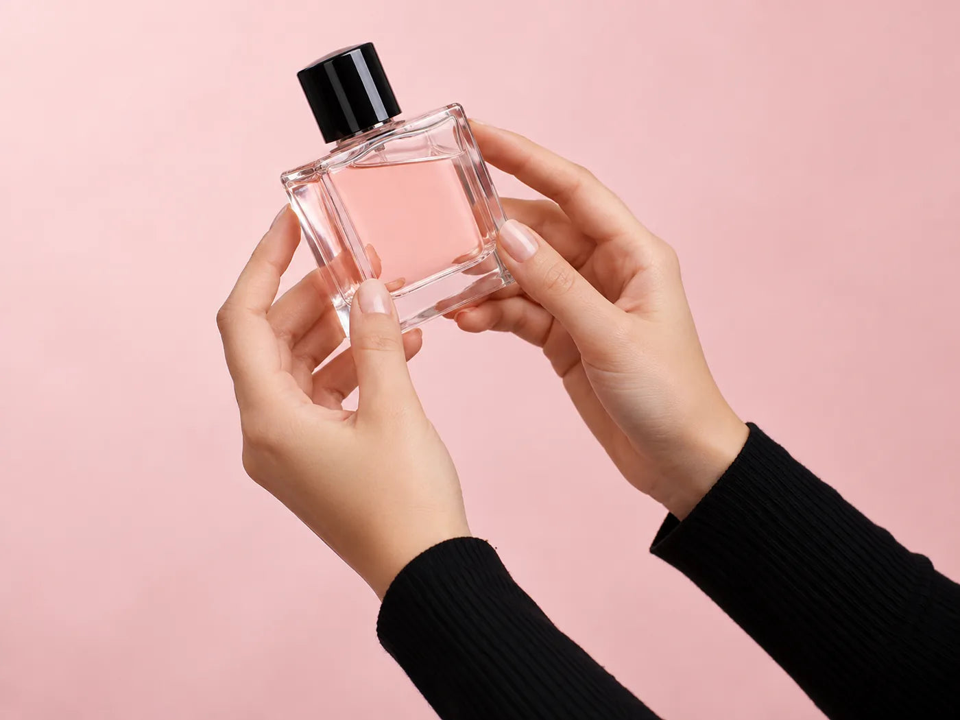 Two hands in black sleeves holding an unlabelled square glass perfume bottle against a blush pink backdrop