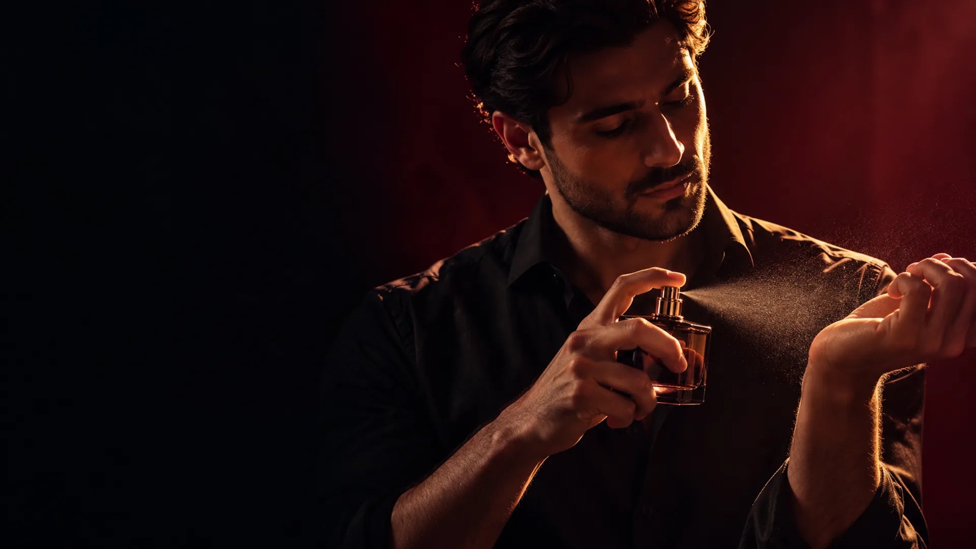 A man in a black shirt spraying perfume onto his wrist against a dark red backdrop